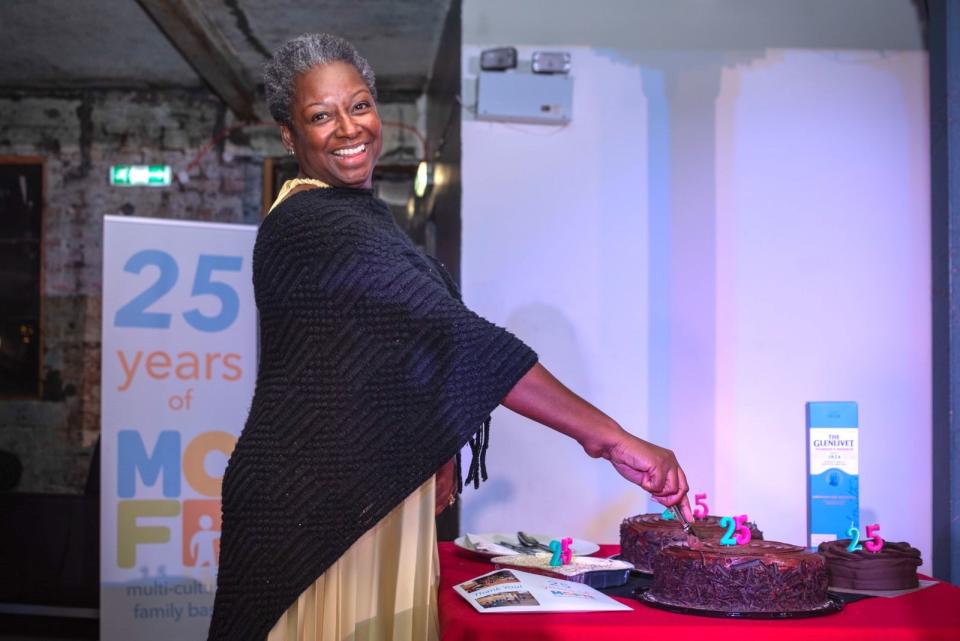Our CEO Ethelinda wearing a yellow dress and black shawl, cutting a cake with 25 number candles. 