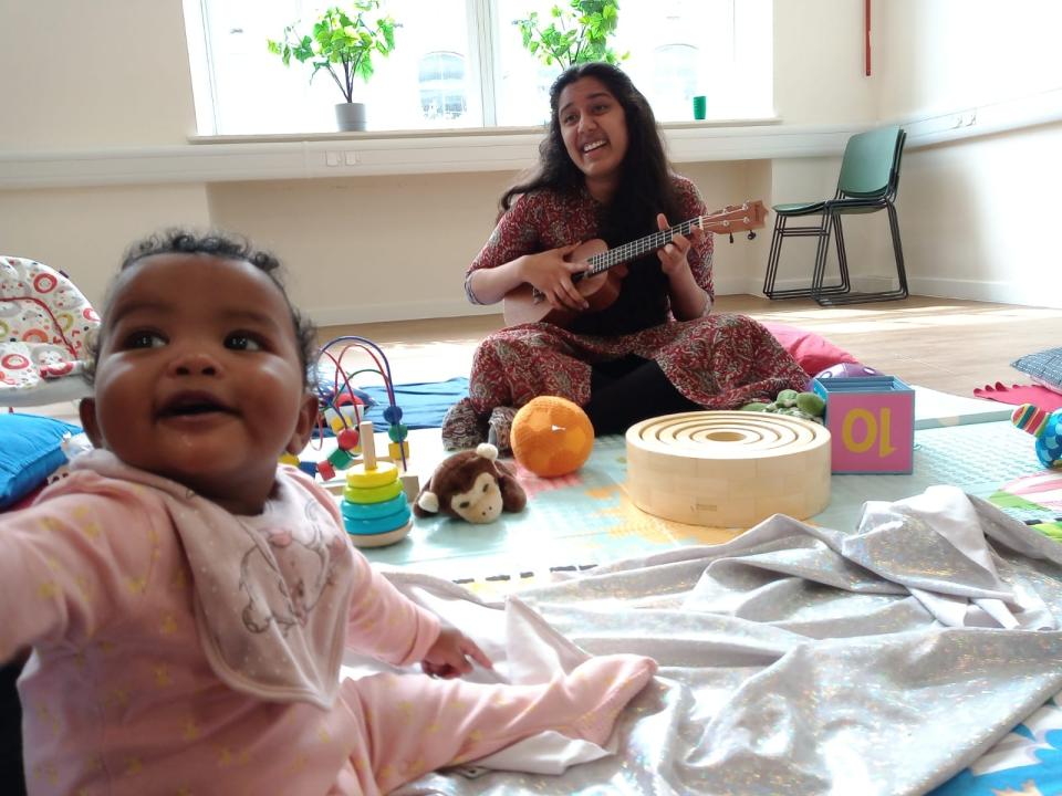 One of our students plays a ukelele during one of the play groups. A baby in the foreground is turned to look at someone behind the camera.