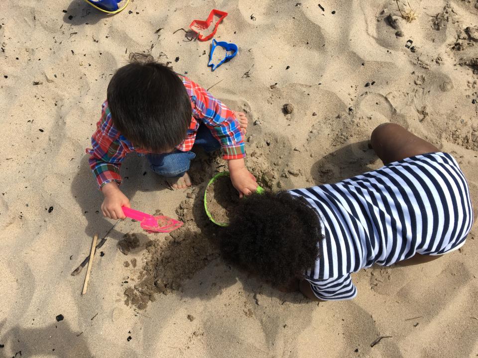 Two toddlers playing with a shovel and pail on the beach. One wears a striped onesie and the other wears a flannel and jeans.