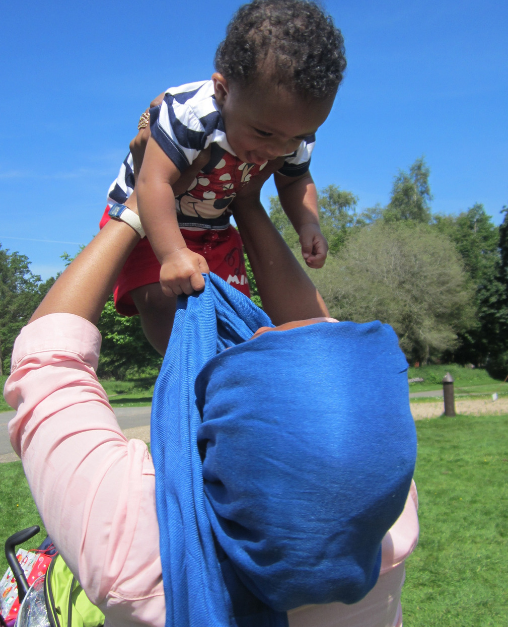 A woman in a blue hijab lifts a baby above her head in an outdoor space. The sky is clear and blue, and the baby is smiling and tugging on her headscarf.