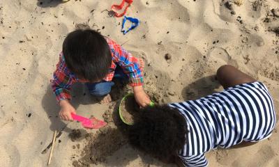 Two toddlers playing with a shovel and pail on the beach. One wears a striped onesie and the other wears a flannel and jeans.